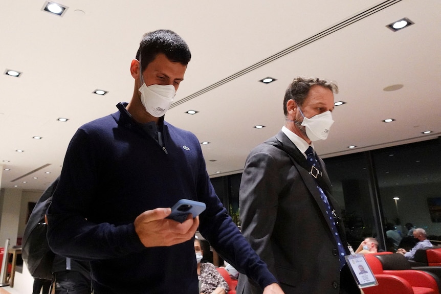 Two men walking, while wearing masks. man on the left looking down at his phone. 