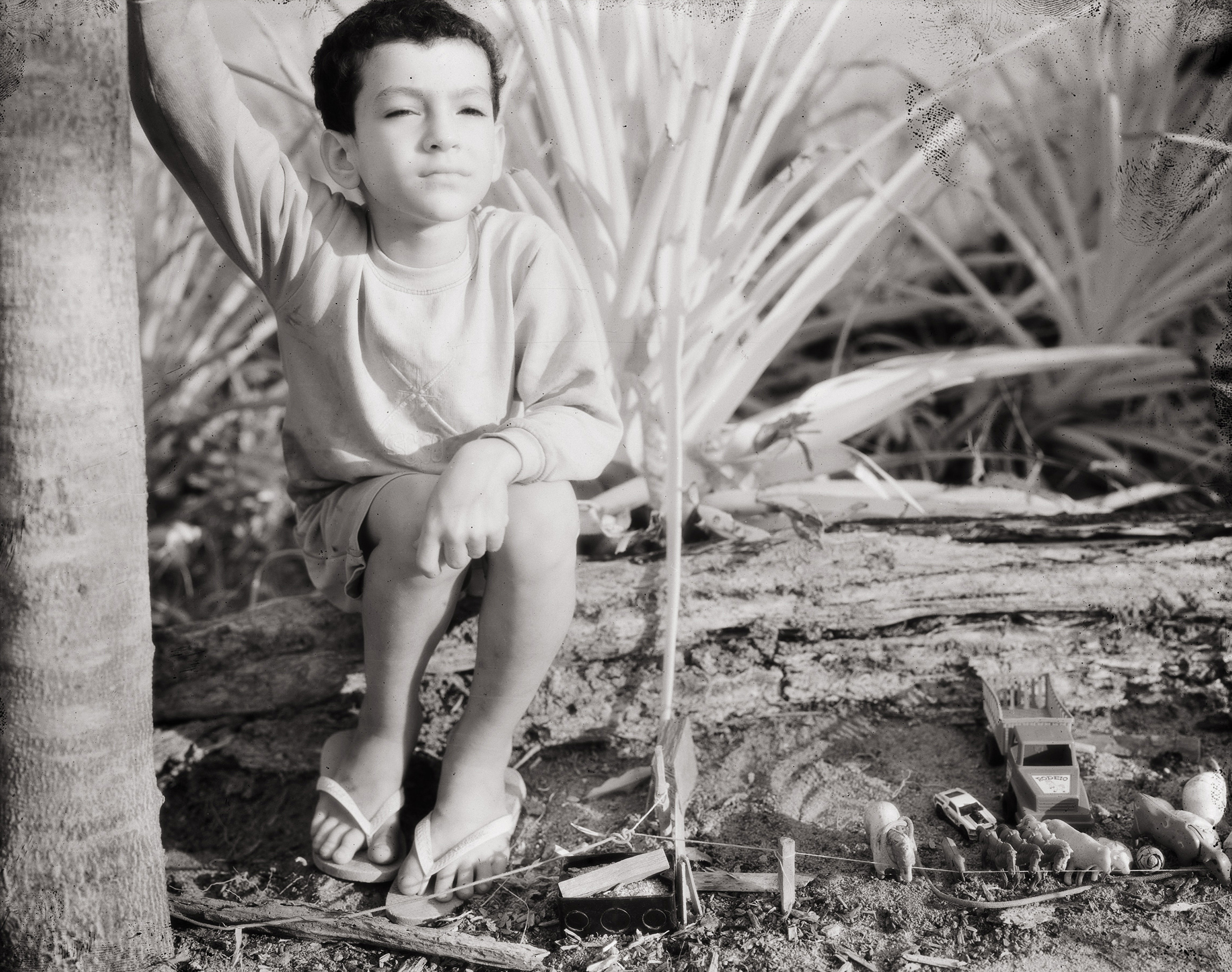 Black and white image of a small boy with dark hair wearing a white long-sleeved shirt and thongs squatting on log in bushland.