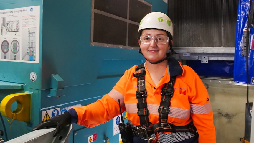 A woman in a high vis shirt, helmet and safety glasses stands in an engine room.