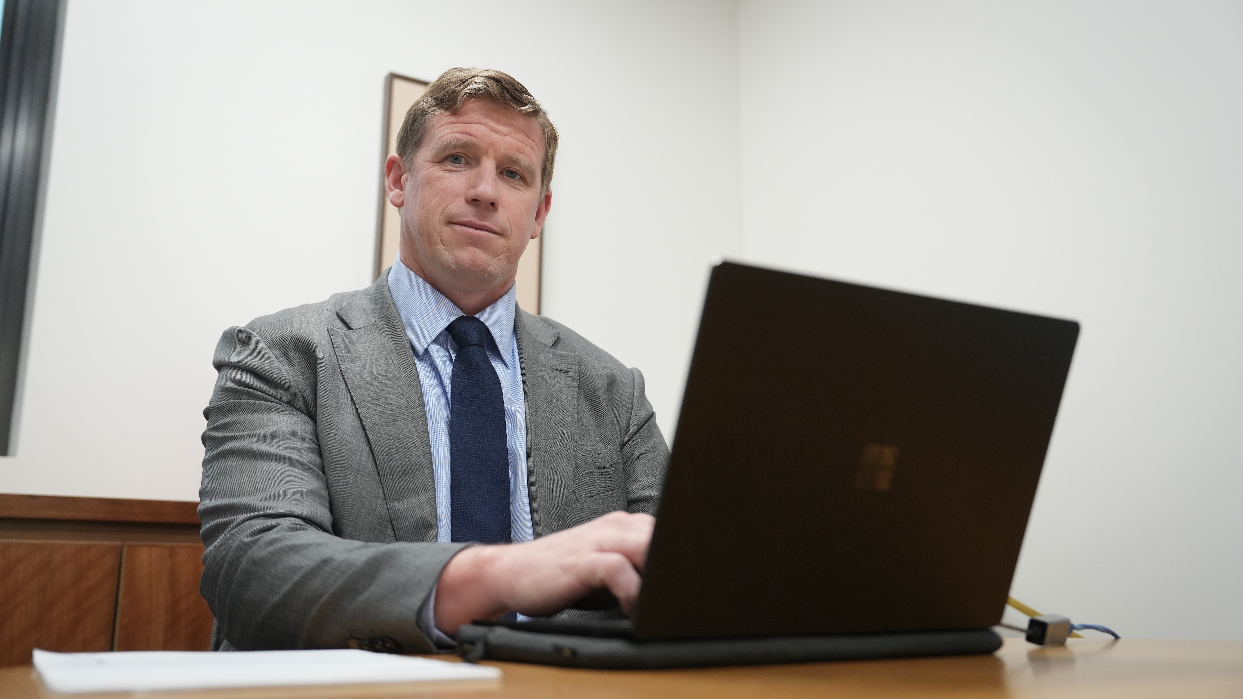 A man with short light hair wearing a grey suit sits at a desk working on a laptop.