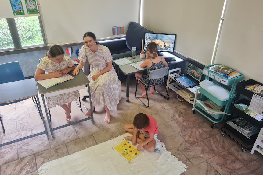 Una donna seduta in un'aula di casa con un bambino per terra, un altro alla scrivania e l'altro al computer.