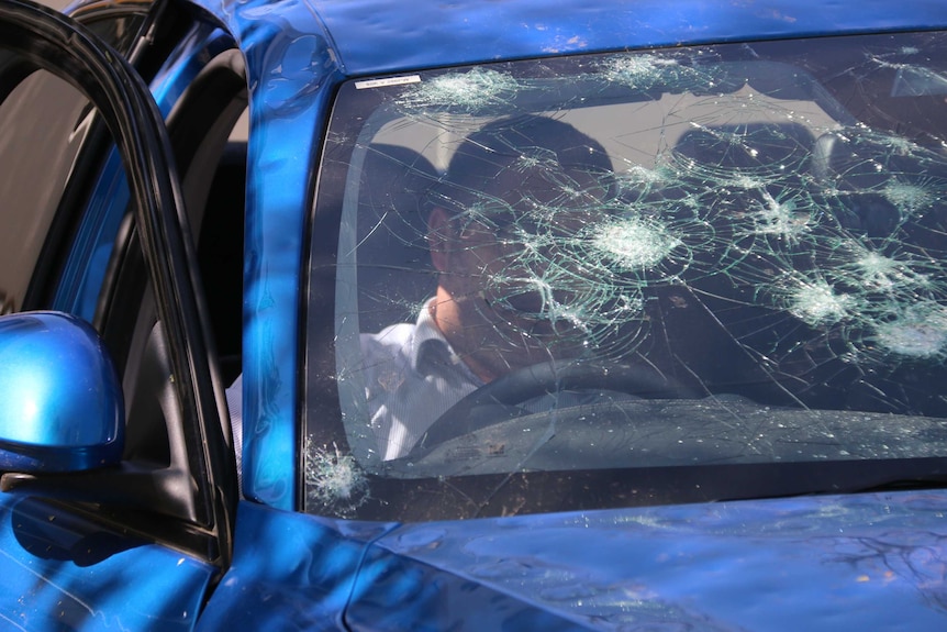 A man sits inside a blue car with a shattered winscreen.