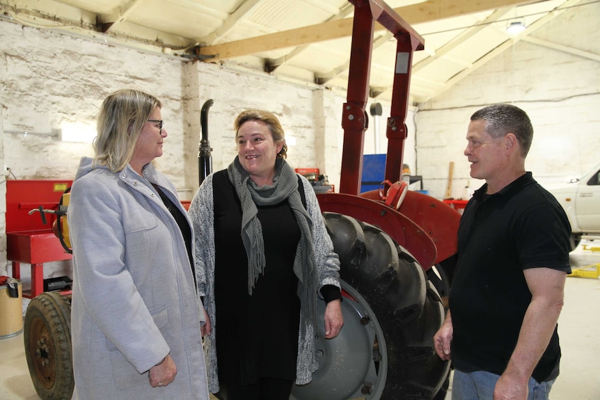 Two women and a man stand in a large, white mechanical workshop.