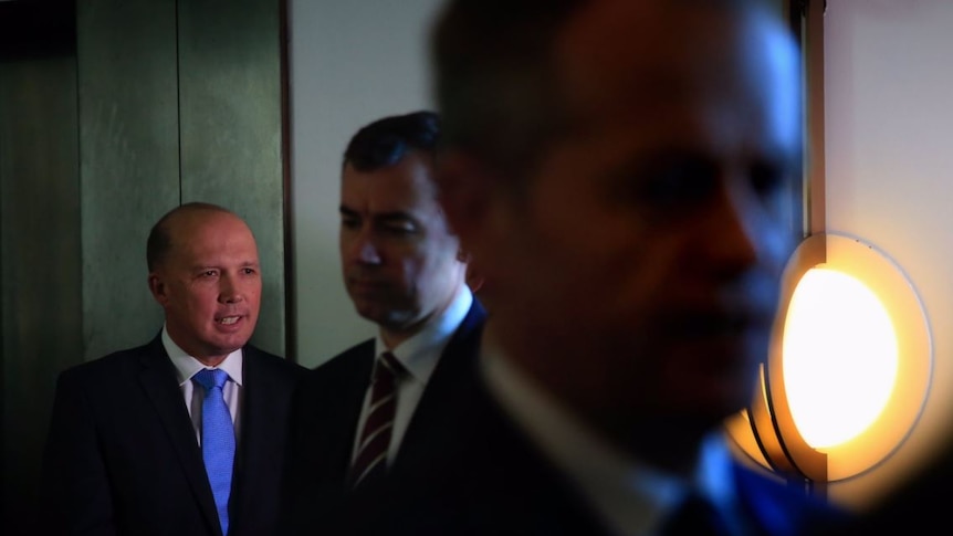 Immigration Minister Peter Dutton stands in the corner in the spotlight as bill shorten and another mp walk away from him