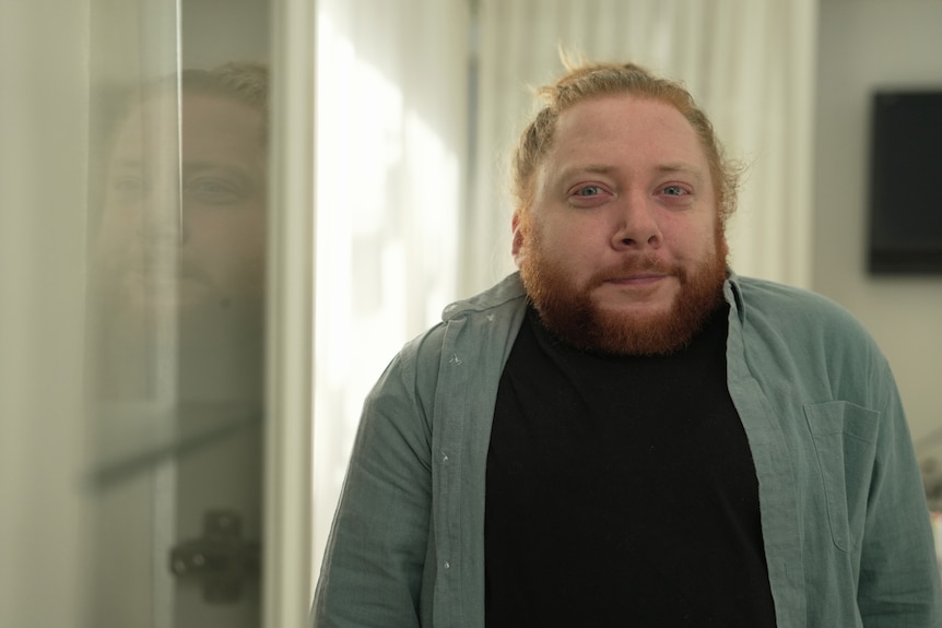 A man from the short statured community in a lounge room. He has red hair and a beard, and is wearing a blue buttoned shirt