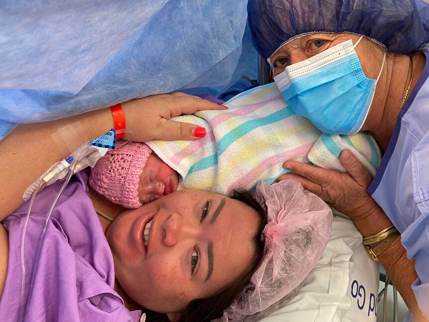 A woman laying down in surgery with a baby in a blanket and beanie on her chest, cuddled by another woman in a a gap and gown.