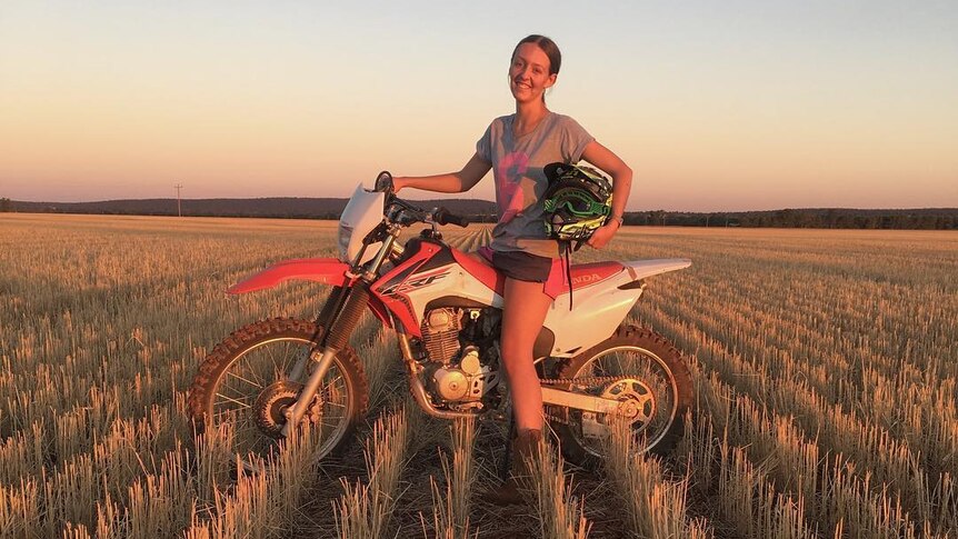 A girl sits on a motorbike in crop stubble at sunset.