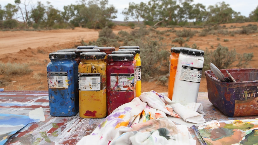 blue, yellow, red, white and orange paint tubs sit on a desk in the red dirt and gum tree outback