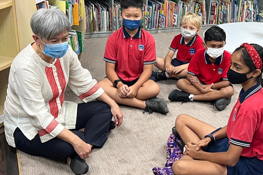 A woman sits cross-legged with school children in a library.