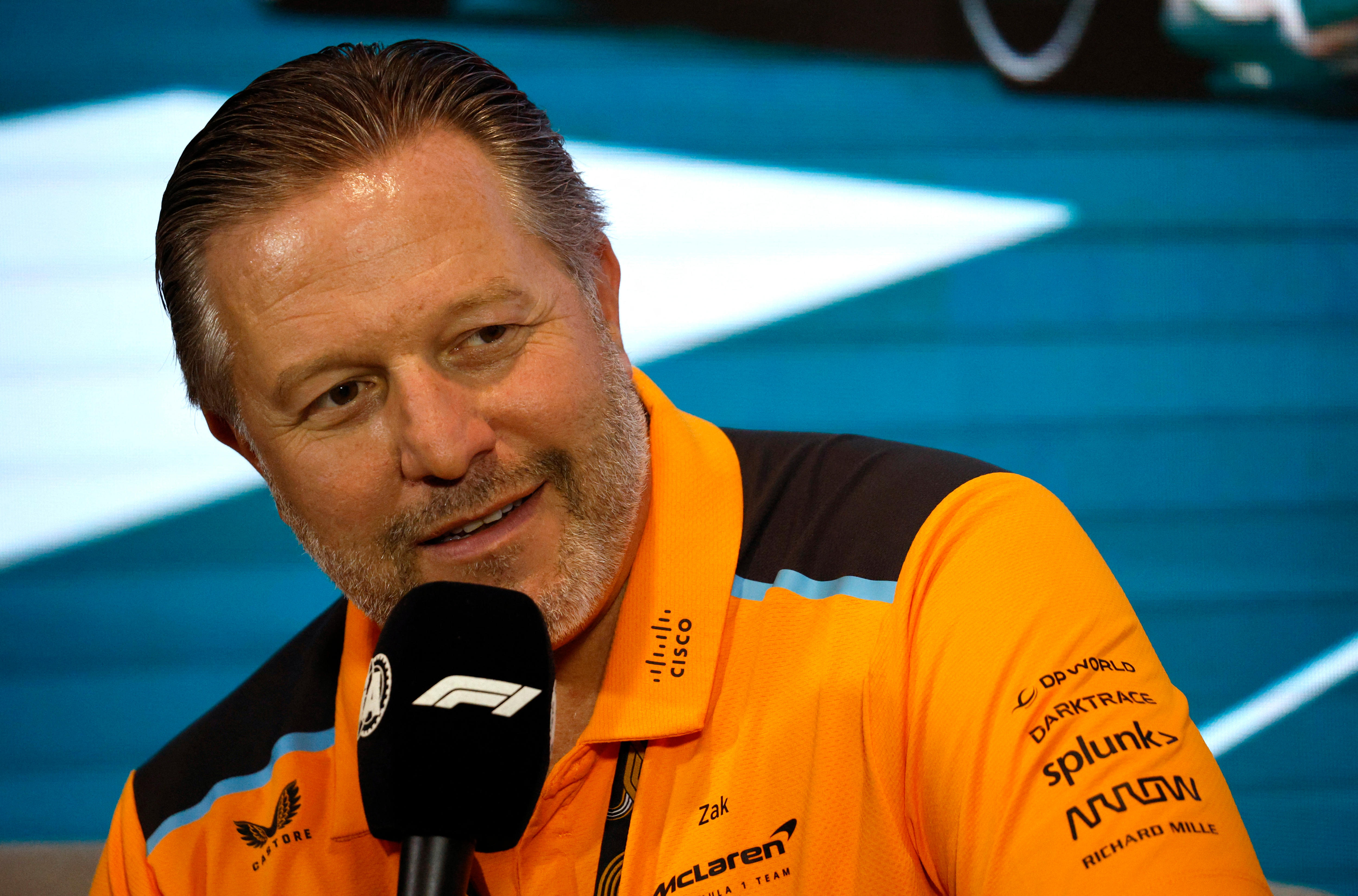 Man in an orange polo shirt, holds a black microphone to his mouth, at a press conference.