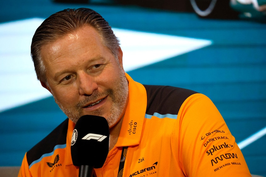 Man in an orange polo shirt, holds a black microphone to his mouth, at a press conference.