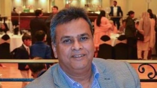 A man wearing a grey suit jacket and blue shirt smiles. He is seated at a table at an indoor function.