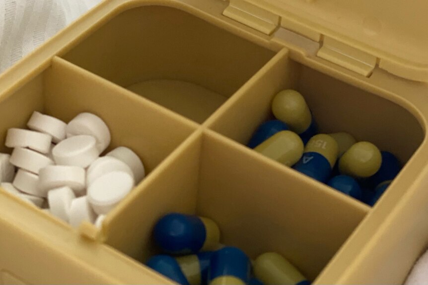 A yellow-beige pill box with four compartments seen with white pills in the bottom left corner, blue and yellow pills on right