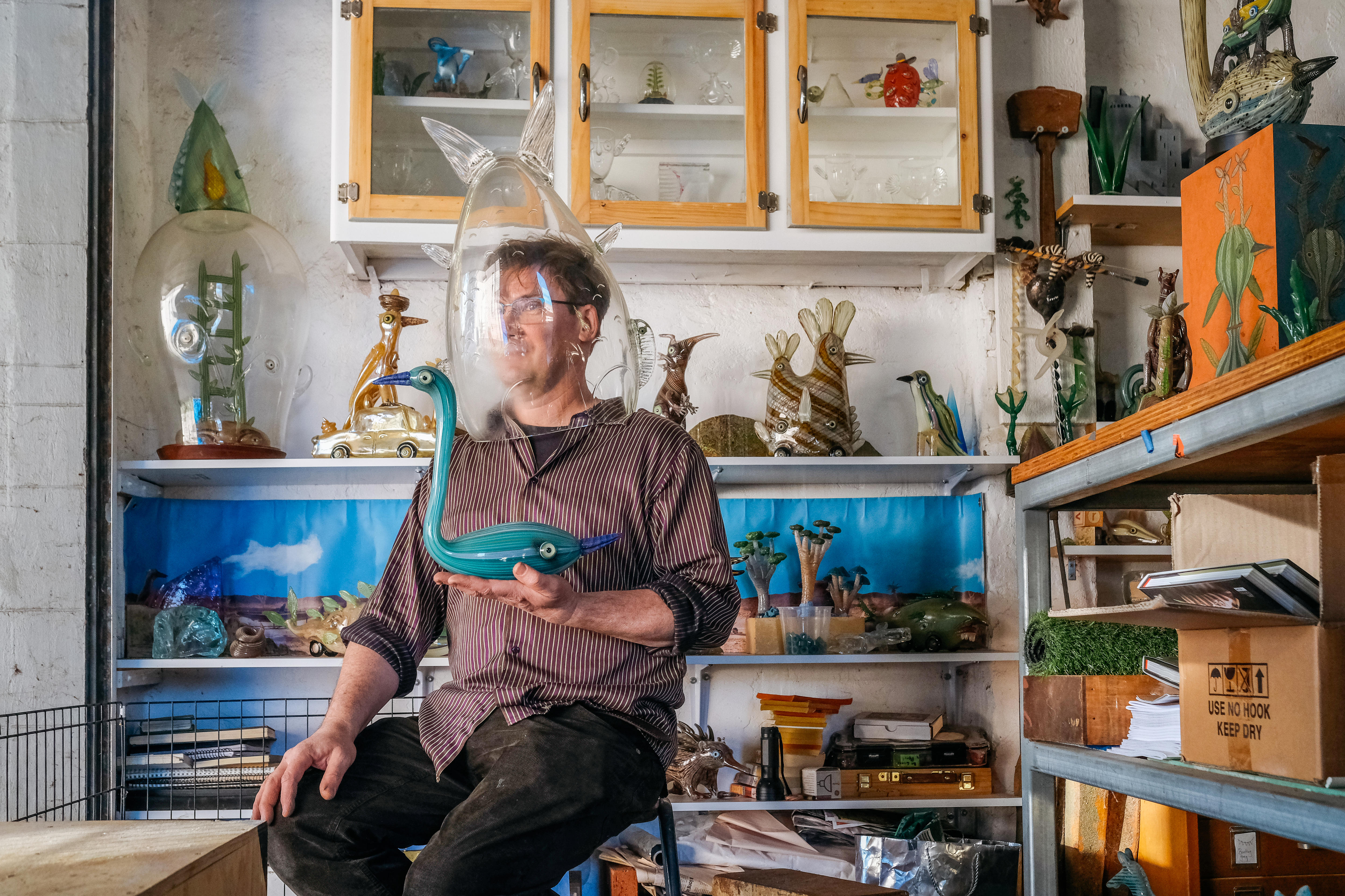 Artist Tom Moore in his studio, surrounded by his colourful glass creations. The middle-aged white artist wears one on his head