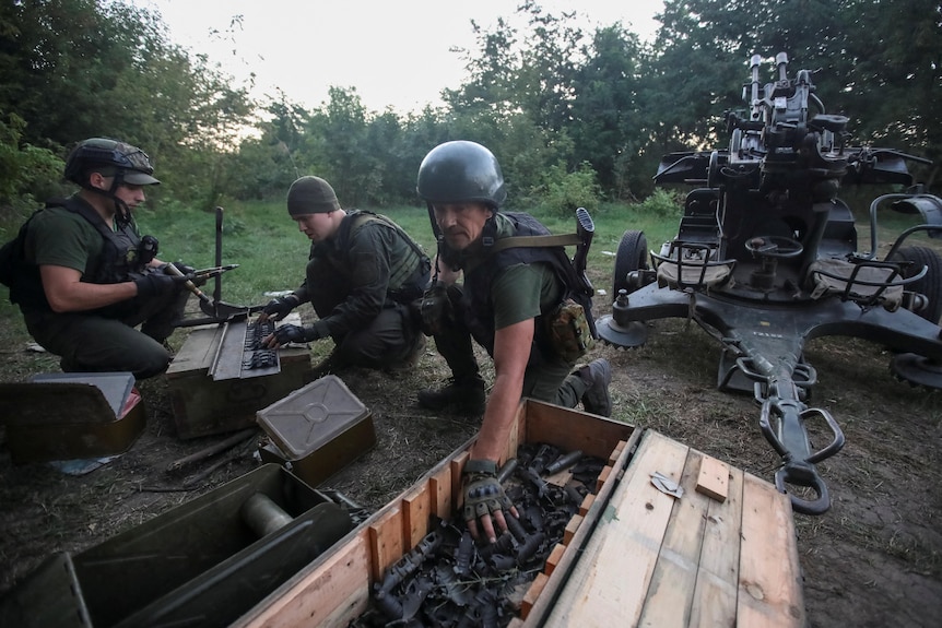 Three Ukrainian soldiers crouch down in a lush green clearing as they rummage in crates next to a large anti-aircraft cannon.
