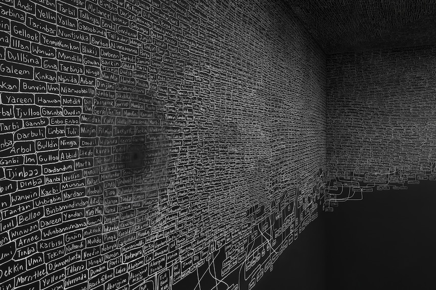 A large black gallery wall is covered in names written in white chalk that are part of a sprawling family tree.