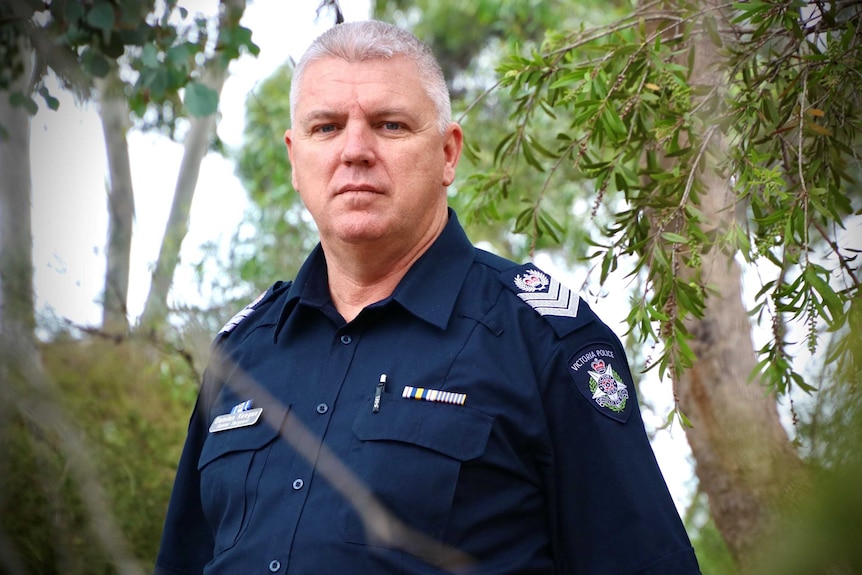 Snr Sgt Damian Keegan stands in uniform looking at country from an area of bushland.