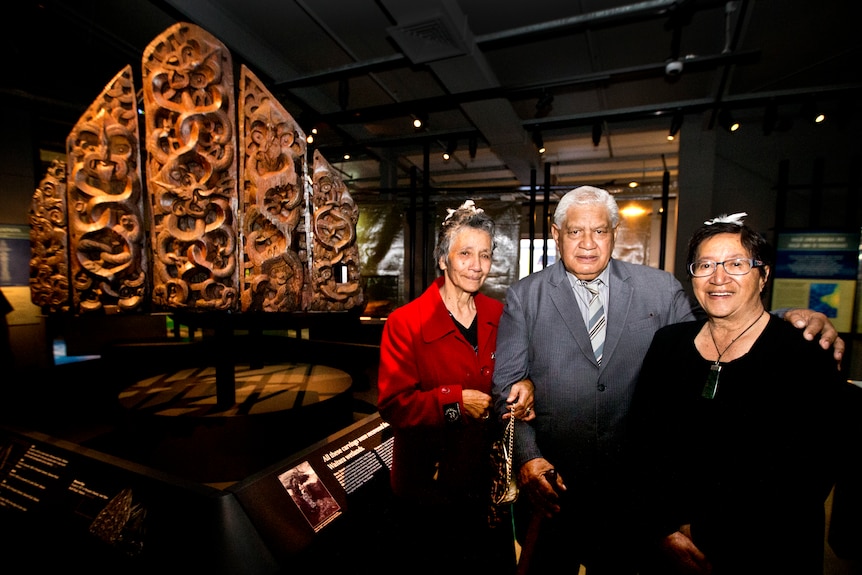 Three people stand in front of carved wooden panels 