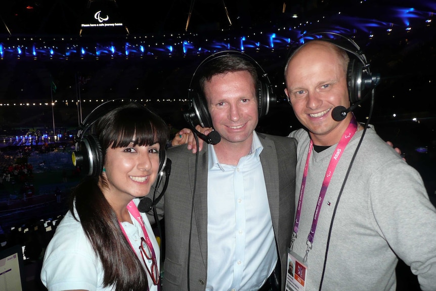A woman and two men arm in arm with headphones on with stadium in background at night.