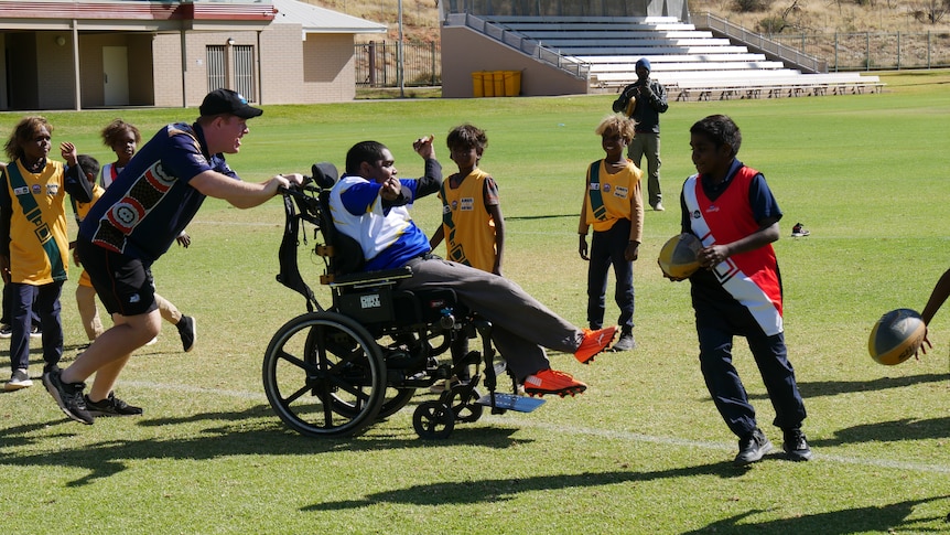 A man in a wheelchair playing Aussie Rules with a group of children.