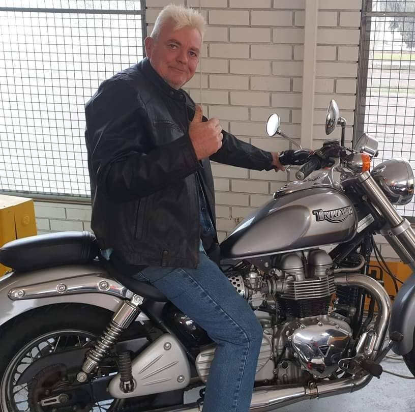 Hombre con cabello gris sentado en una motocicleta sonriendo y mostrando los pulgares hacia arriba 