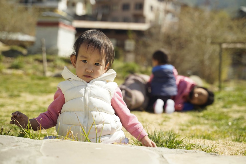 A toddler looks serious while sitting on the grass with her two sisters in the background playing on the grass