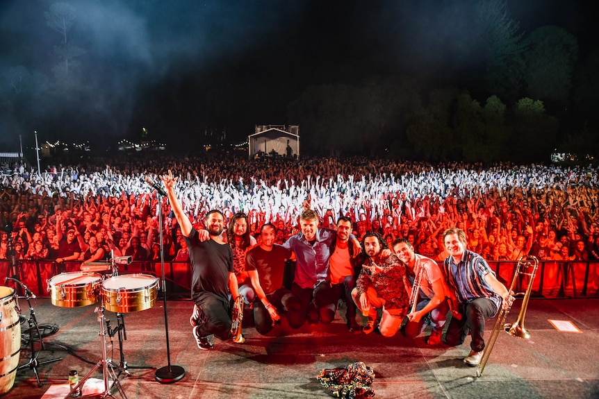 Before a huge crowd under a dark sky, eight members of the Cat Empire squat facing away from the audience to smile for a photo.