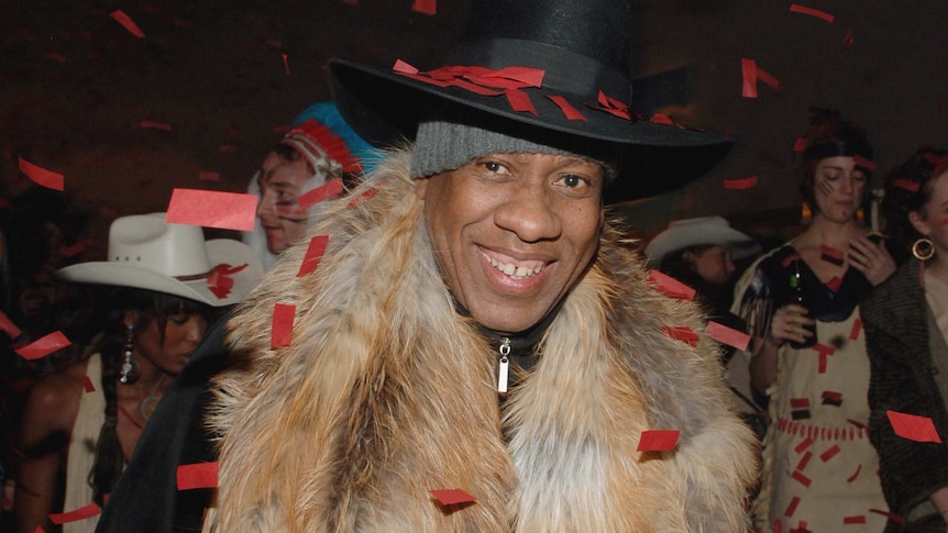 Andre Leon Talley wears a long fur shawl and a black cowboy-style hat as he smiles walking through a burst of red confetti.