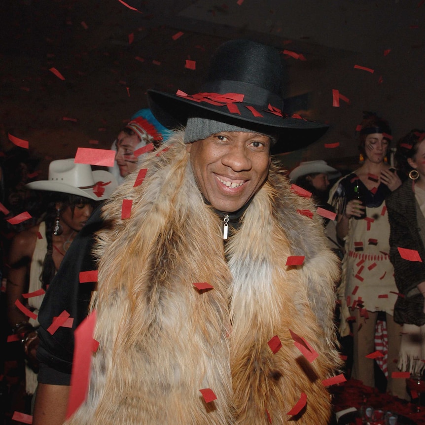 Andre Leon Talley wears a long fur shawl and a black cowboy-style hat as he smiles walking through a burst of red confetti.