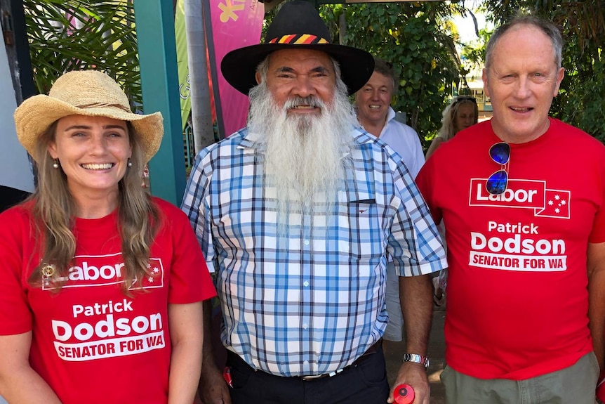 A mid shot showing a smiling Pat Dodson posing for a photo in a checked shirt flanked by two Labor supporters in red shirts.