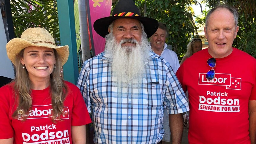 A mid shot showing a smiling Pat Dodson posing for a photo in a checked shirt flanked by two Labor supporters in red shirts.