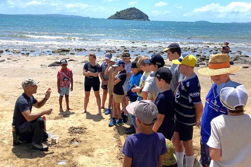 An instructor wearing a black shirt, pants and army cap talking to children on a sandy beach with an island in the background.