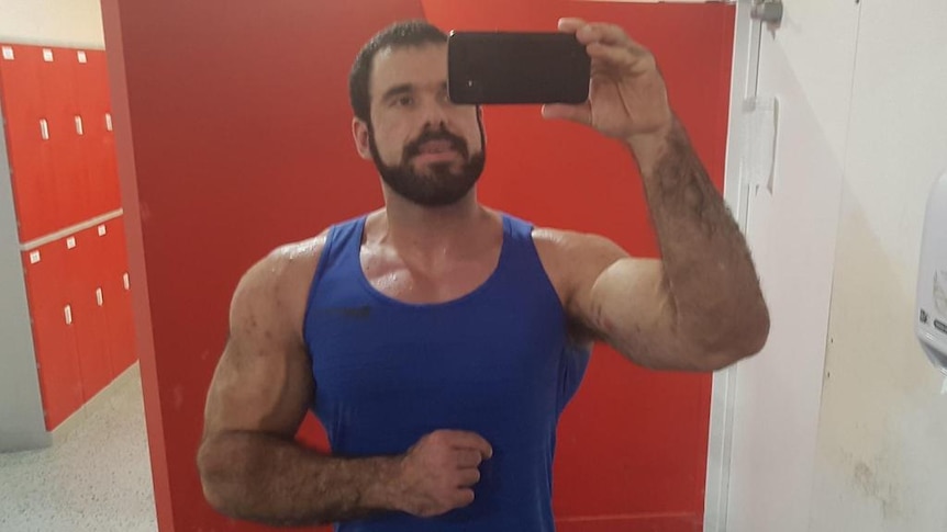 A man with dark hair and facial hair wearing a blue singlet flexes his biceps in a mirror selfie