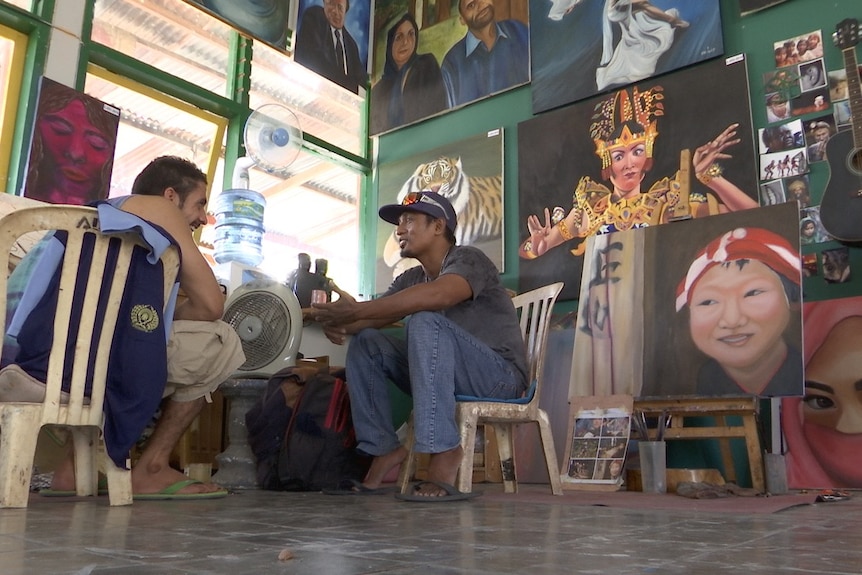 Two men sit in plastic chairs chatting, surrounded by artwork