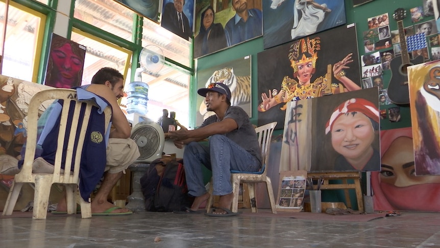 Two men sit in plastic chairs chatting, surrounded by artwork