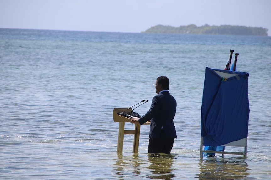 Мужчина в костюме стоит у кафедры по бедра в морской воде, произнося речь.  
