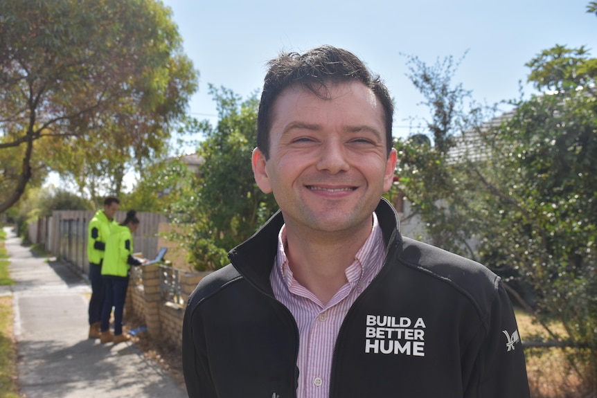 Naim Kurt wears a black jacket that says "build a better Hume" and stands near two workers on a street footpath.