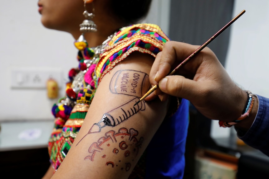 A woman in colourful dress has a COVID-19 vaccine syringe painted on her upper arm. 