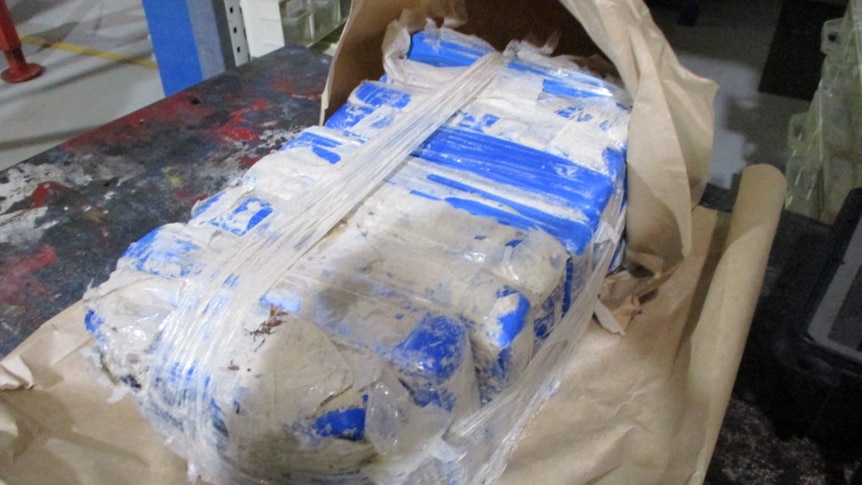 Ten blue plastic packages dusted with white powder are taped together, in a paper bag, on a bench.