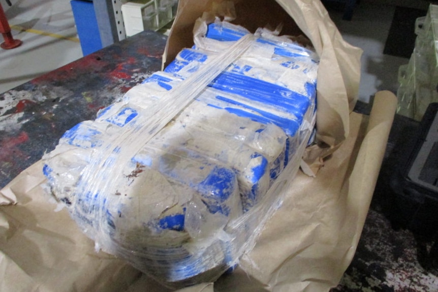 Ten blue plastic packages dusted with white powder are taped together, in a paper bag, on a bench.