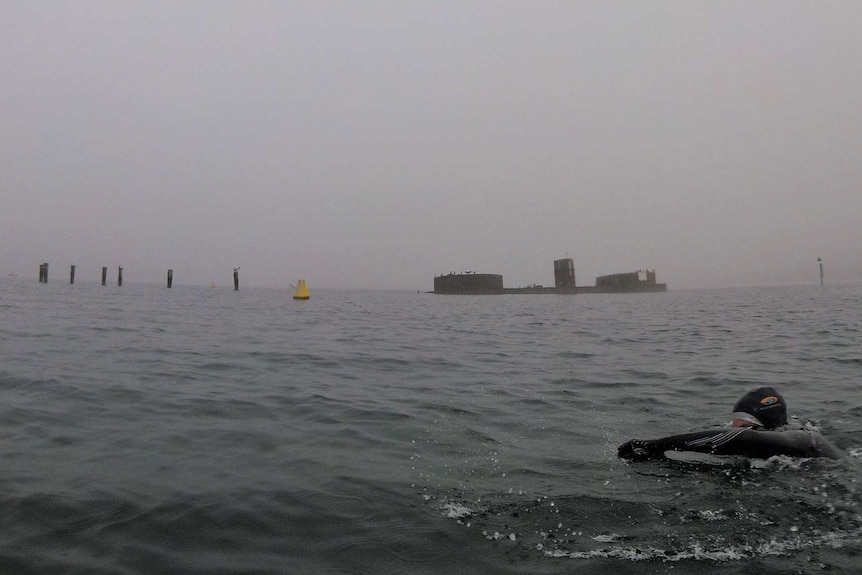 A man in a wetsuit swims in the ocean, with the wreck of an old submarine in the background