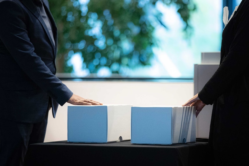 Two white boxes are seen on a table. Two men in suits stand by each box, gently touching it.