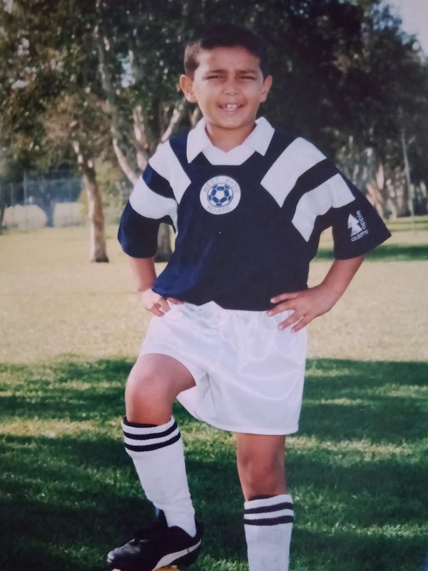 Childhood photo of smiling Mohamad Ikraam Bahram dressed in team uniform for soccer, with his foot on a football on a field.