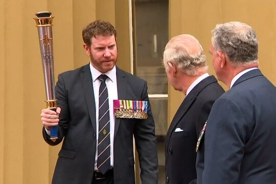 A man in a suit decorated with medals holds a large torch while speaking to King Charles.