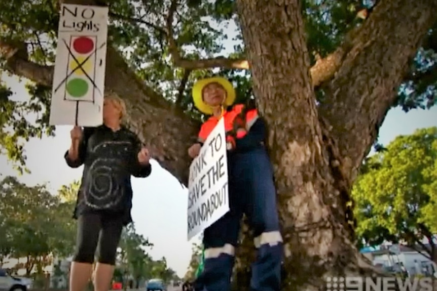 Grainy TV screenshot of two people with placards protesting to save a roundabout. The man is chained to the tree.
