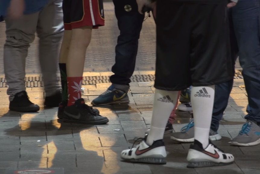 Several young men, photographed from the legs down, standing on a city pavement wearing street gear.