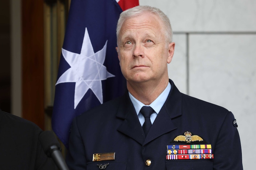 Binskin is in uniform, looking skyward, standing in front of an Australian flag.