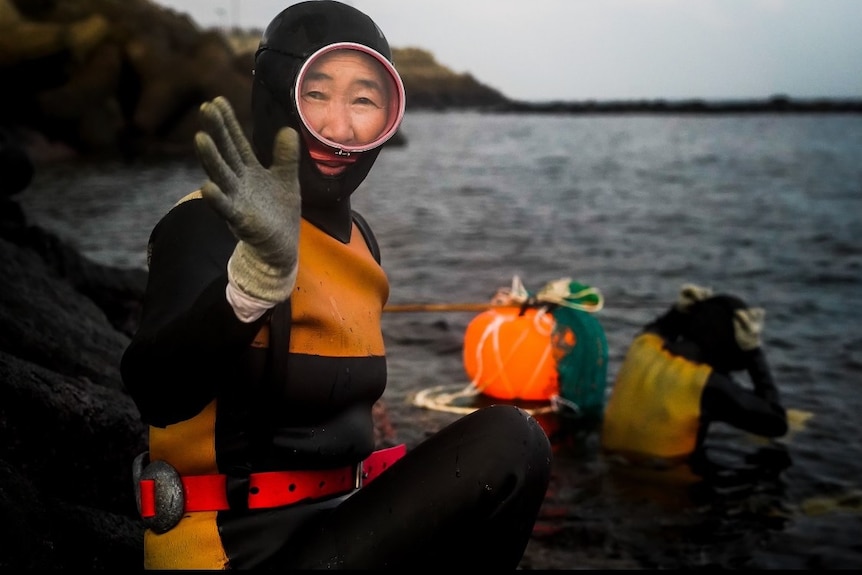 A woman wearing a diving suit, goggles and gloves waves as she sits by the sea.