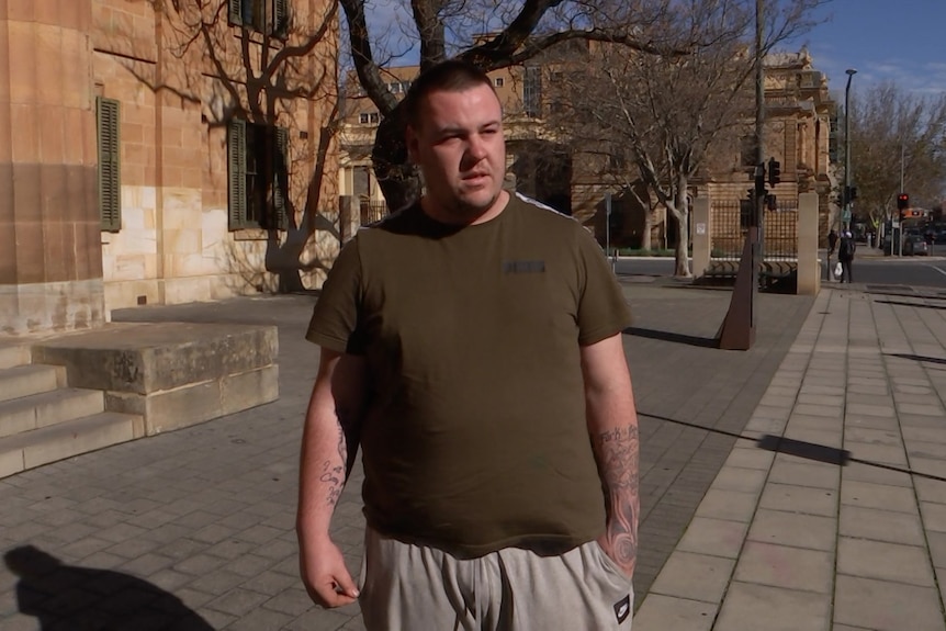 A man wearing a brown T-shirt and grey trackpants in front of a sandstone building on a city street 