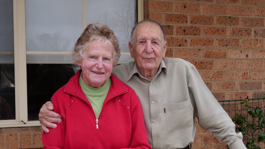 An older couple smile at the camera with their arms around one another from the front balcony of a brick residence